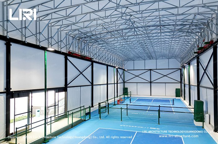 Blue pickleball courts beneath a tall aluminum roof frame and white sidewalls
