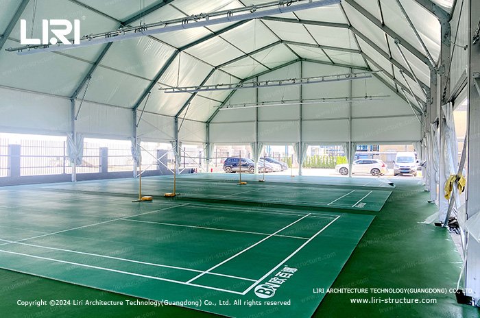 Green badminton courts inside a tent with raised side curtains and daylight entering along the sides